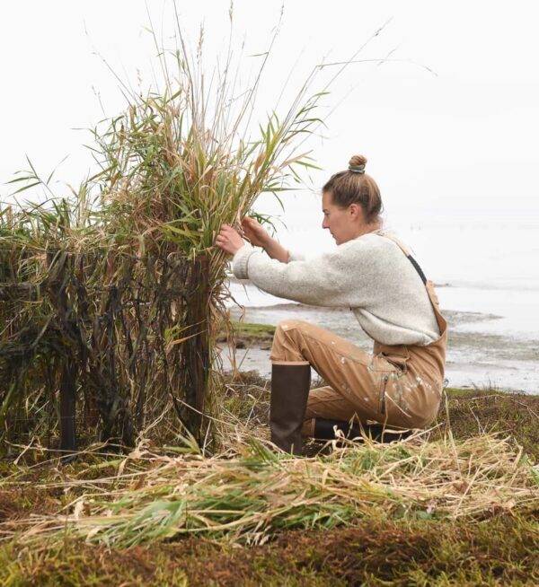 A woman works in front of a body of water, sitting on top of hay wearing boots and a pair of brown overalls made by Dovetail Workwear.