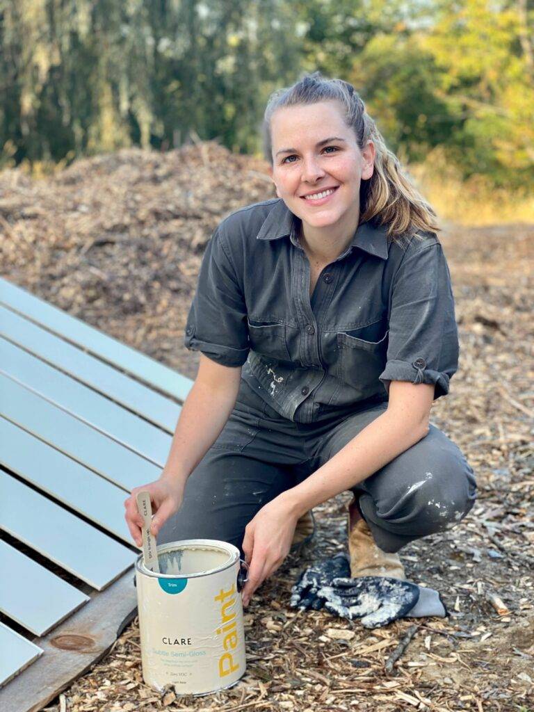 Bea wears a painting coverall while smiling to camera and stiring a container of paint.