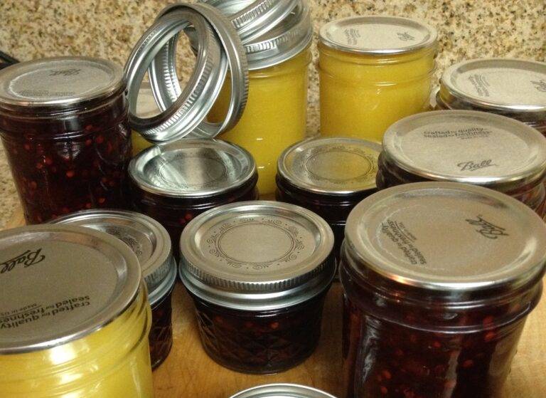 Assorted clear glass canning jars, some filled with creamy yellow curd and others with dark red jam, sit on a cutting board with the rings close by to screw on top of the lids.